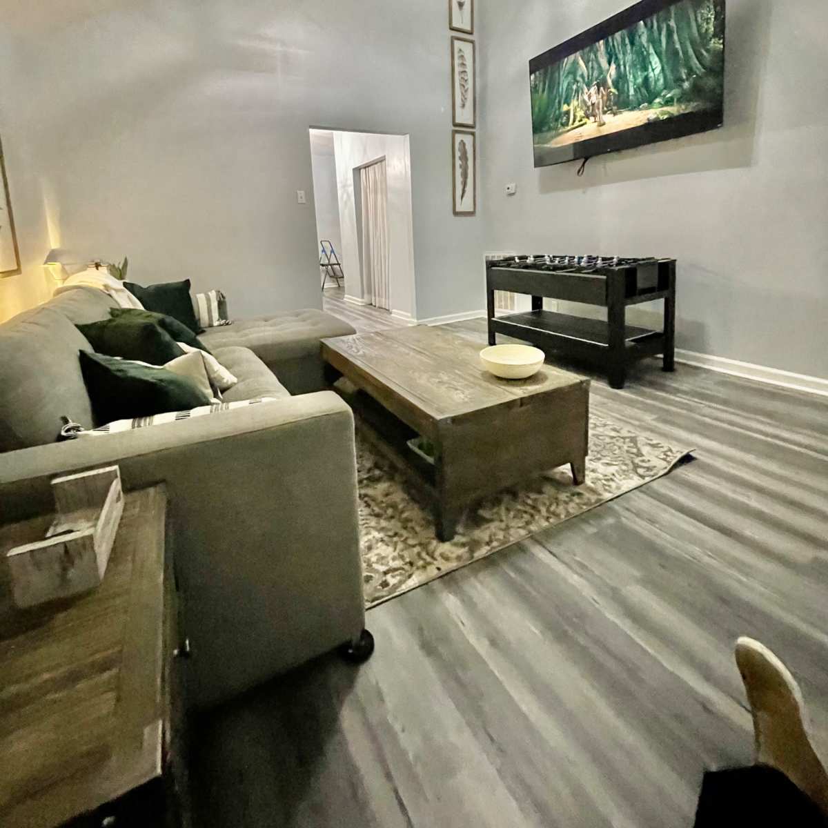 The living room features a gray sectional sofa, a wooden coffee table, and a television mounted on the wall, with light-colored walls and hardwood flooring.