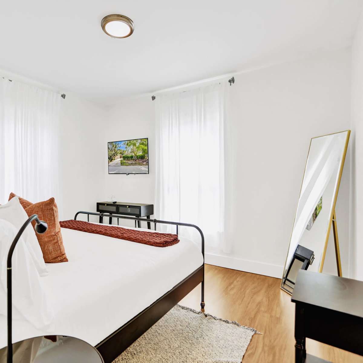 A bright bedroom featuring a bed with white bedding, a large mirror, a dresser, and a television mounted on the wall.