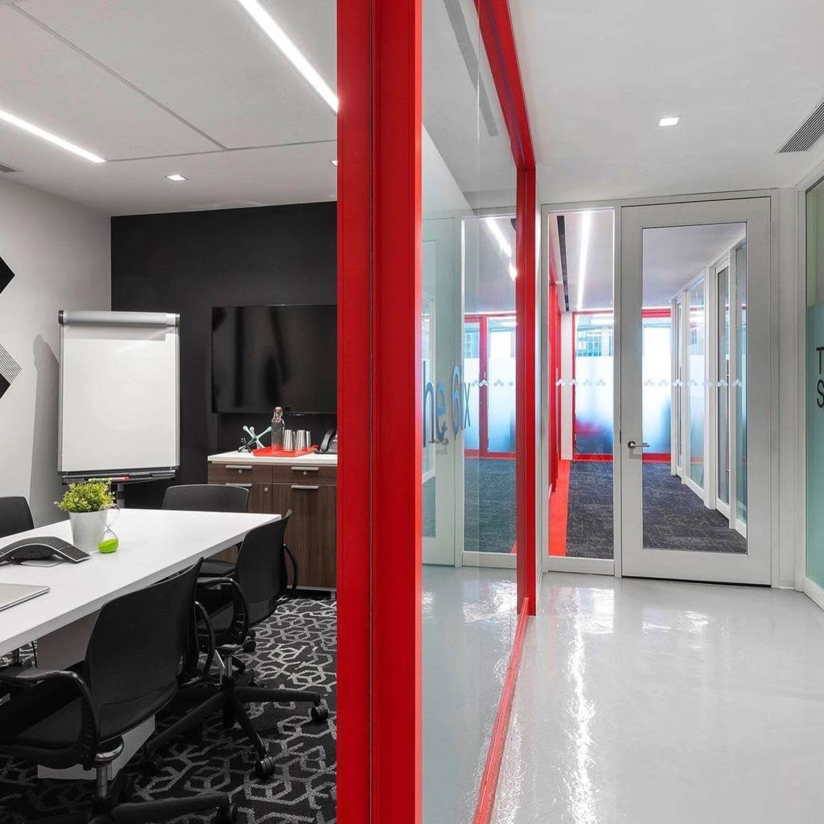 A modern meeting room with a white table and black chairs, featuring a large screen and a glass wall leading to a hallway with red accents.