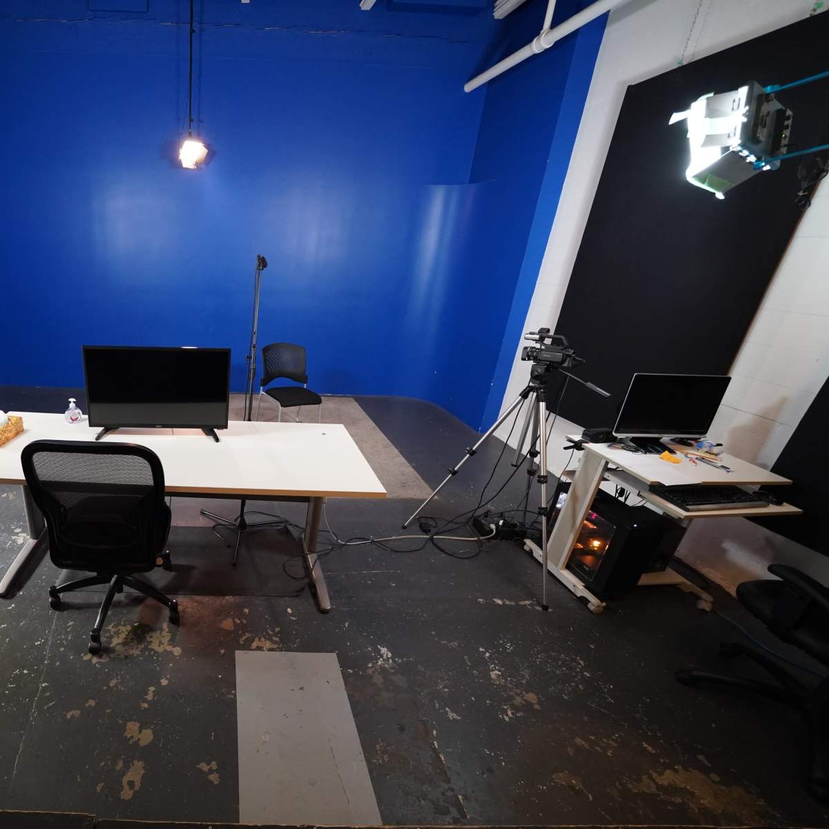 A sparsely furnished studio with a blue wall, a desk with a monitor, a chair, and lighting equipment set up for recording.