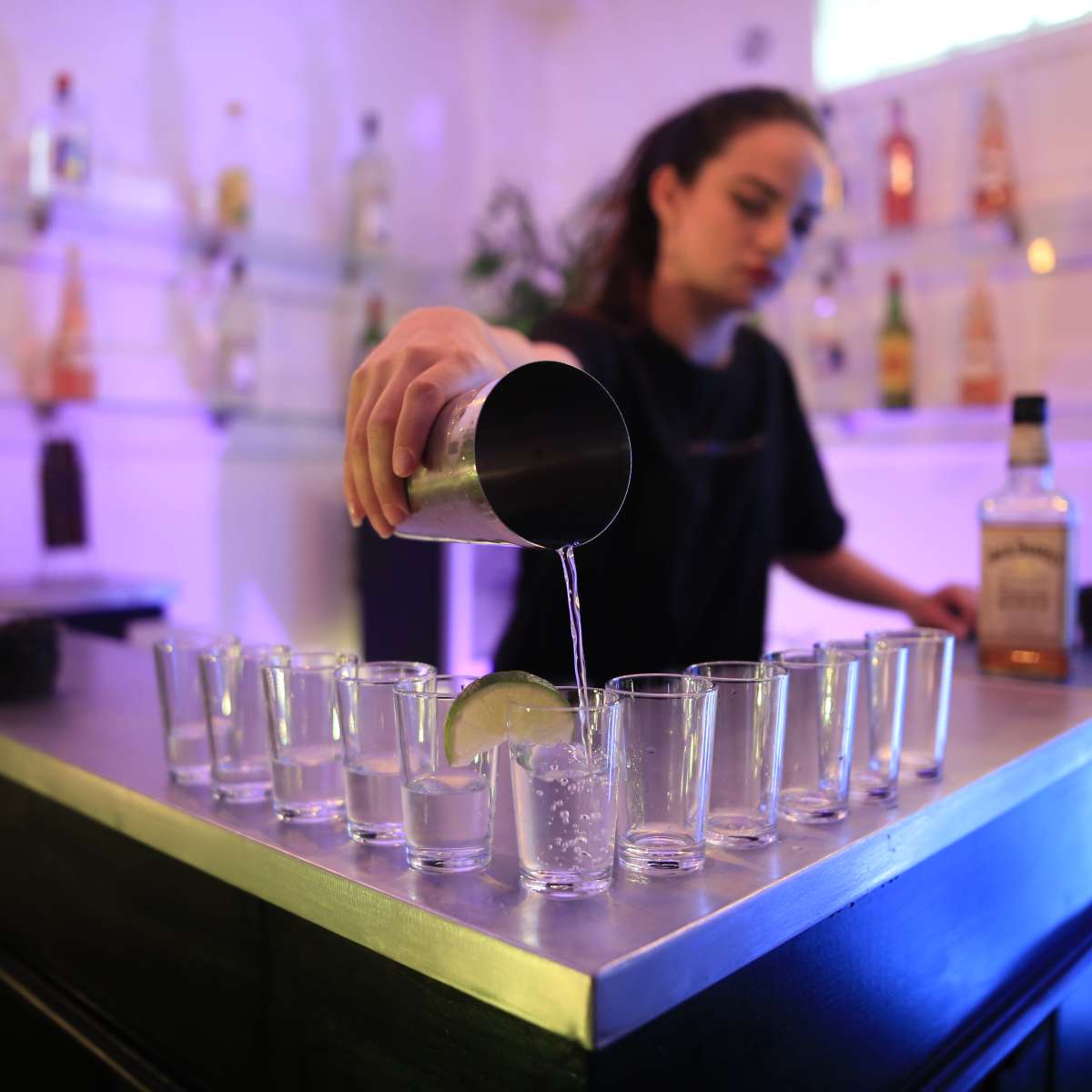 A bartender is pouring a drink into a shot glass lined up on a bar counter.