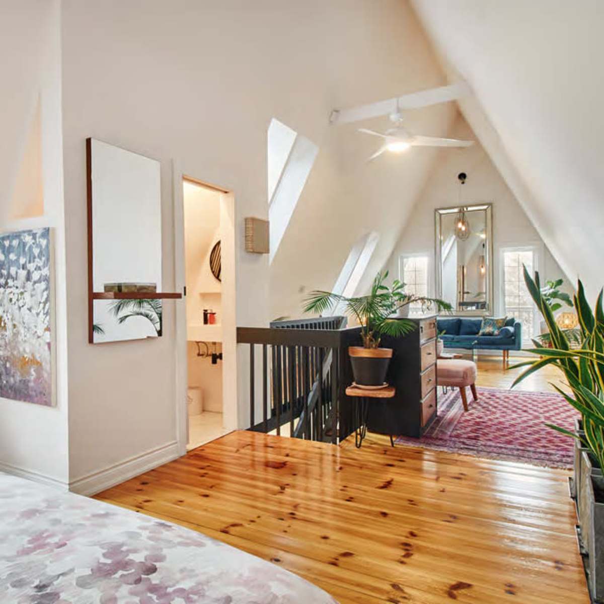 A spacious, well-lit room with angled ceilings, a wooden floor, and decorative plants, leading to a sitting area with a mirror and artwork.