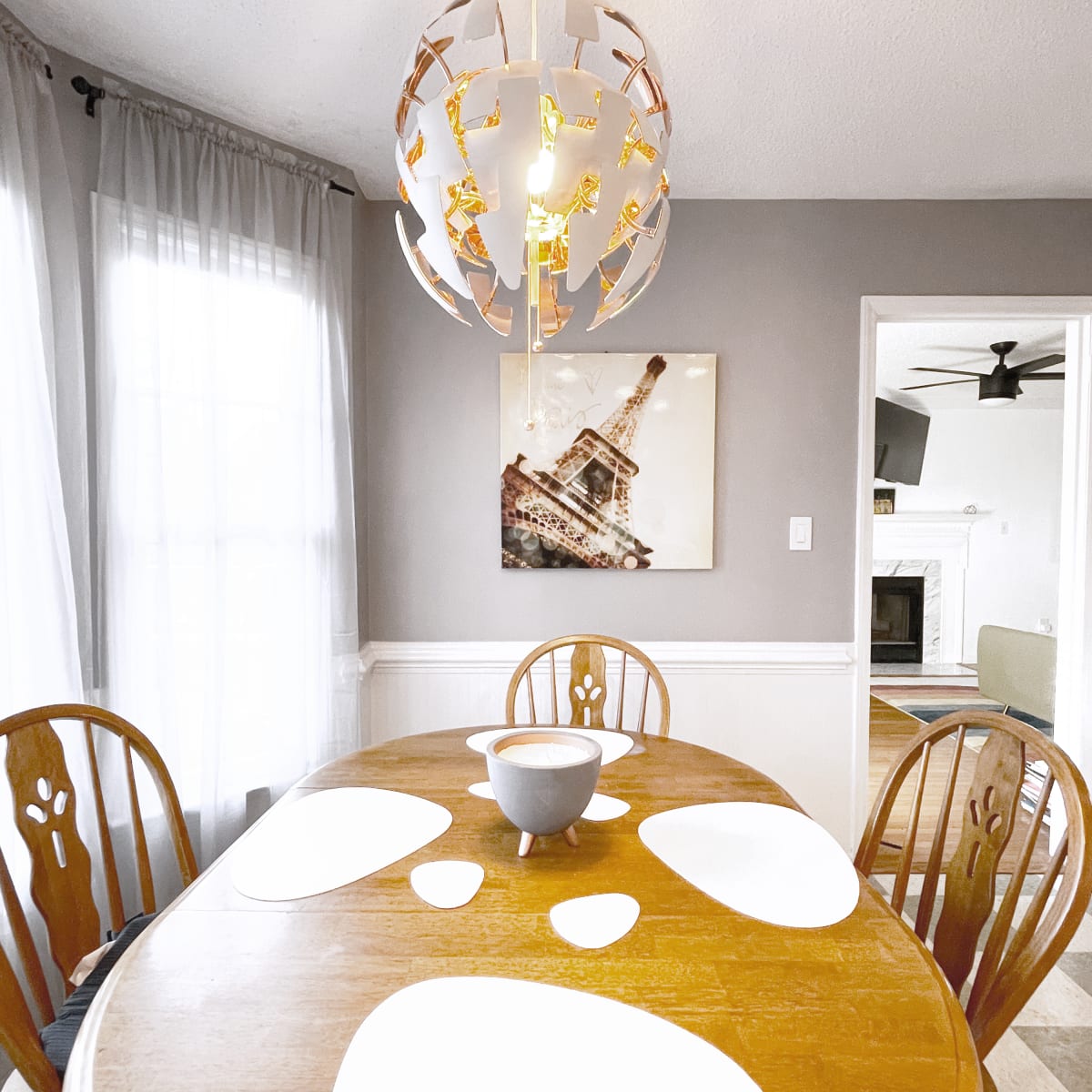 A wooden dining table set with white placemats and a bowl sits beneath a decorative light fixture in a room featuring a wall art piece of the Eiffel Tower.