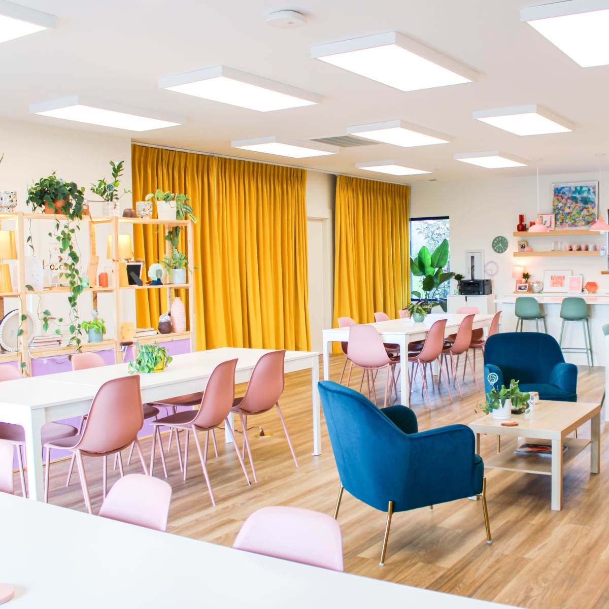 A modern office space with multiple work tables, colorful chairs, a kitchenette area, and large windows with yellow curtains.