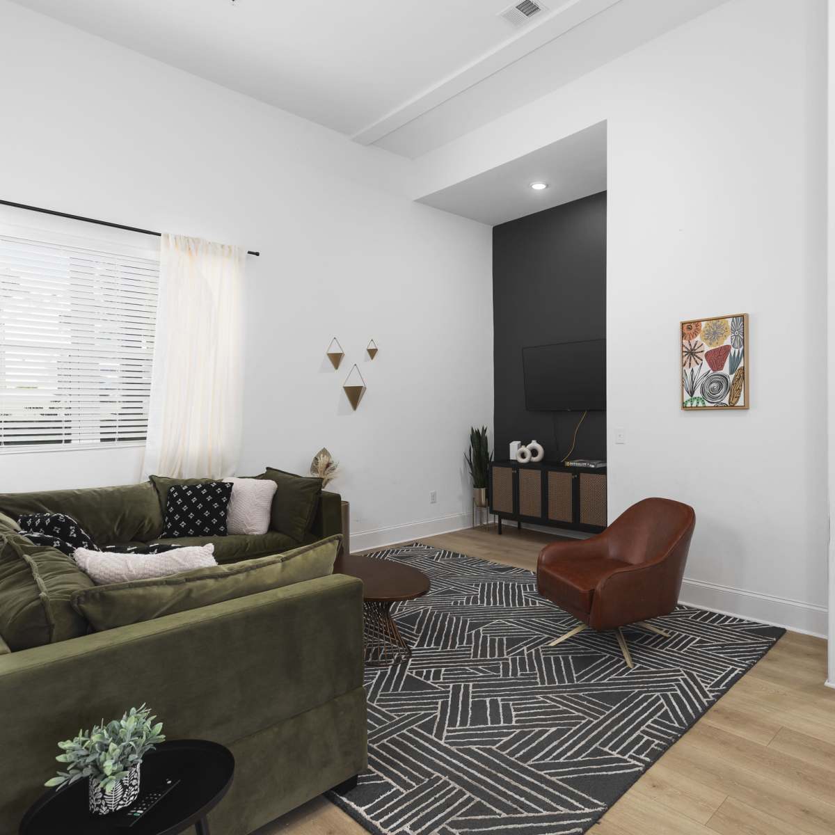 The living room features a green sectional sofa, a round coffee table, and a large television mounted on a dark accent wall, with a hallway leading to other rooms in the background.