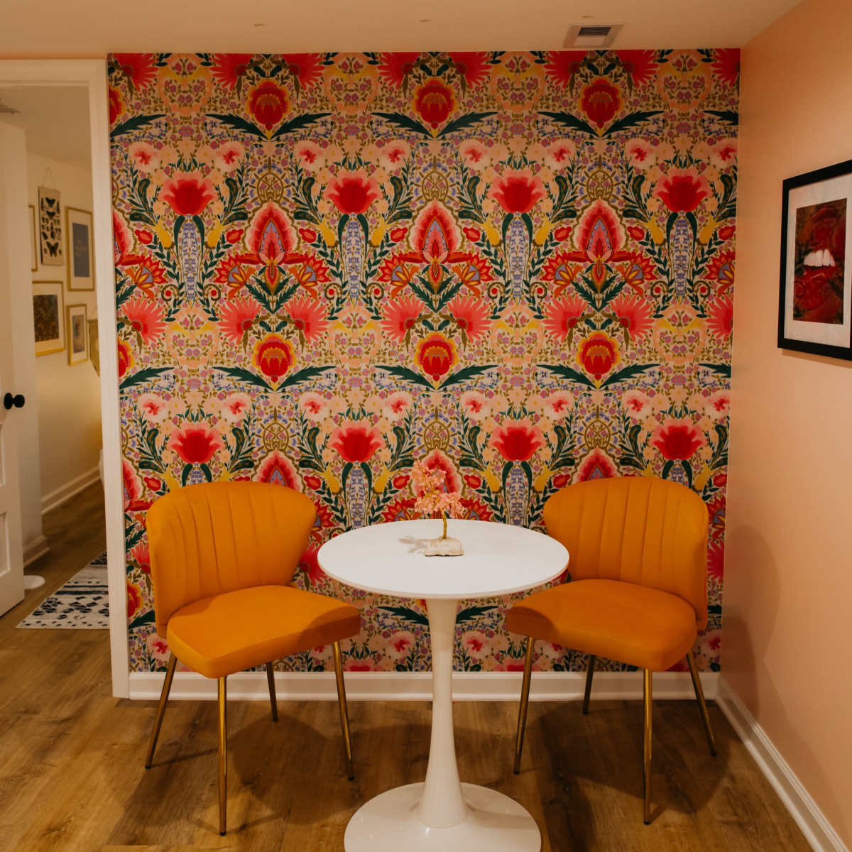 A small dining area featuring a round white table and two orange chairs against a vibrant floral patterned wallpaper.