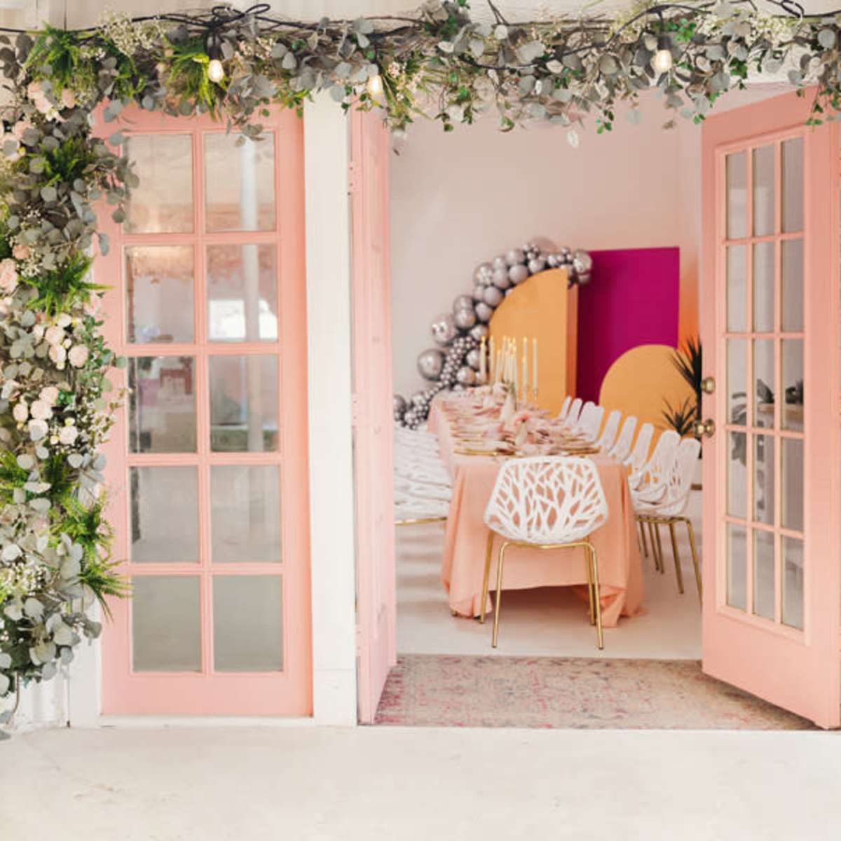 A beautifully decorated entrance with pink double doors framed by greenery, leading into a bright event space with a dining table set for a gathering.