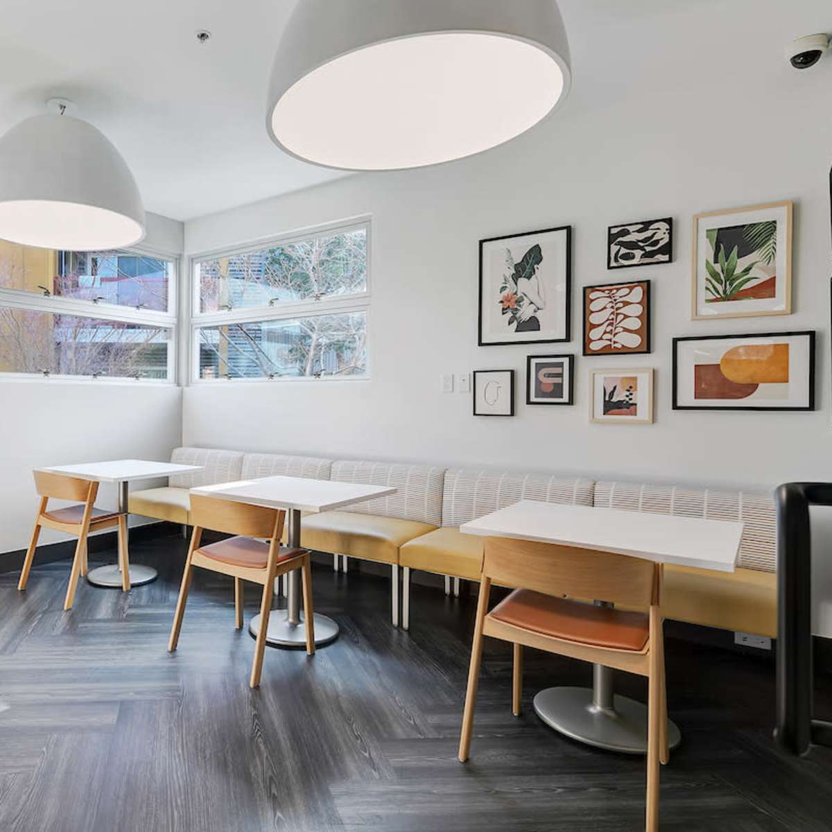 A bright, minimalist café interior featuring wooden tables, chairs, and large pendant lights, alongside a wall adorned with framed artwork.