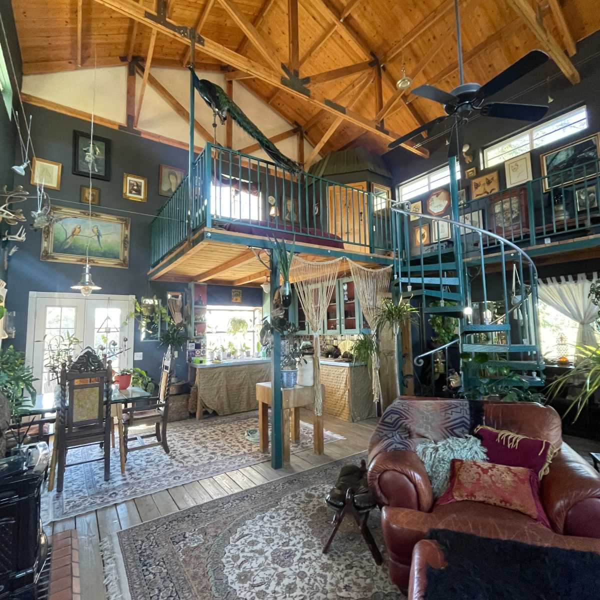 A spacious, rustic living area with high wooden ceilings, a spiral staircase leading to a loft, and various plants and artworks adorning the walls.