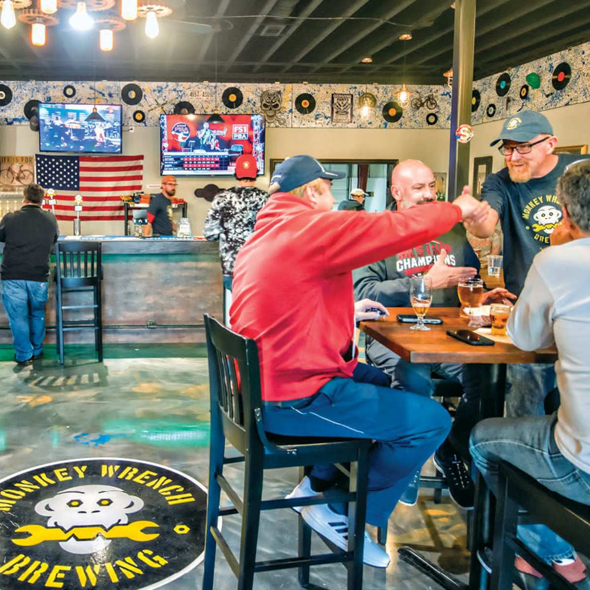 A brewery interior features patrons socializing at tables and a bar, with televisions displaying sports and a large American flag on the wall.