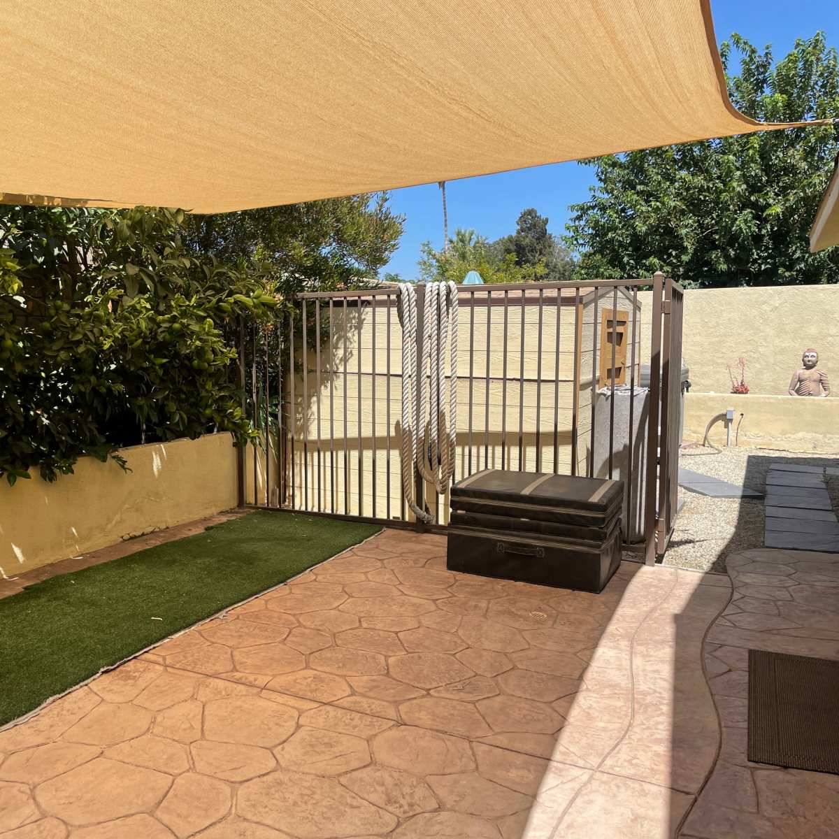 A patio area with a shaded section, artificial grass, a gated enclosure, and a storage box against a wall.