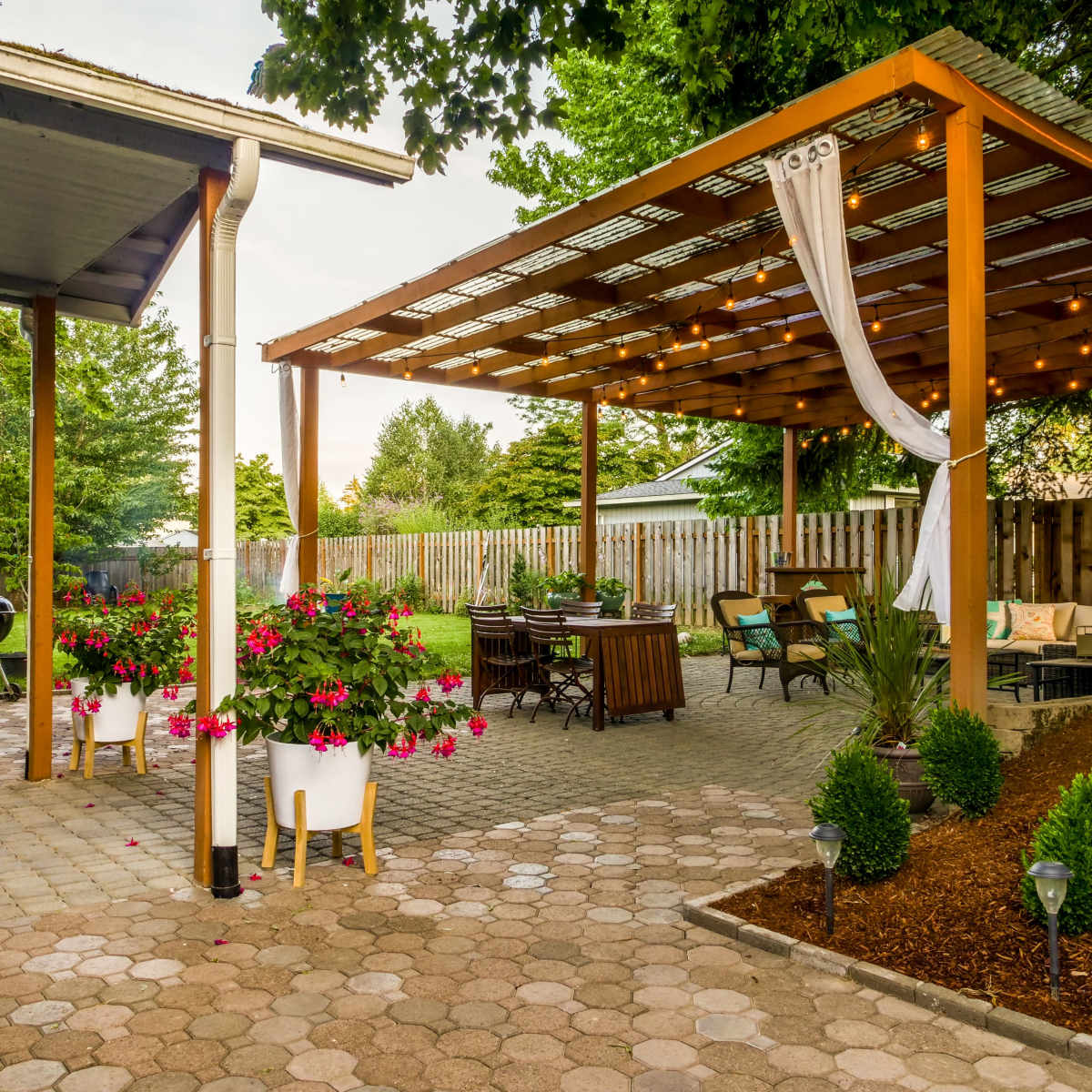 A backyard patio with a wooden pergola, outdoor seating, hanging lights, and flower pots next to a paved walkway.