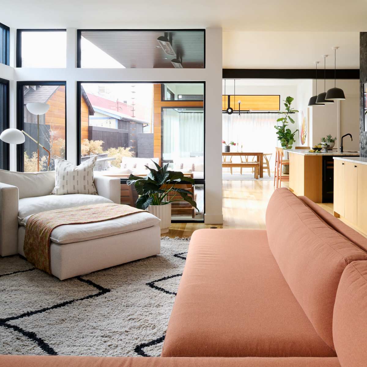 A modern living room with large windows, a sectional sofa, a lounge chair, and an open layout leading to a dining area and kitchen.