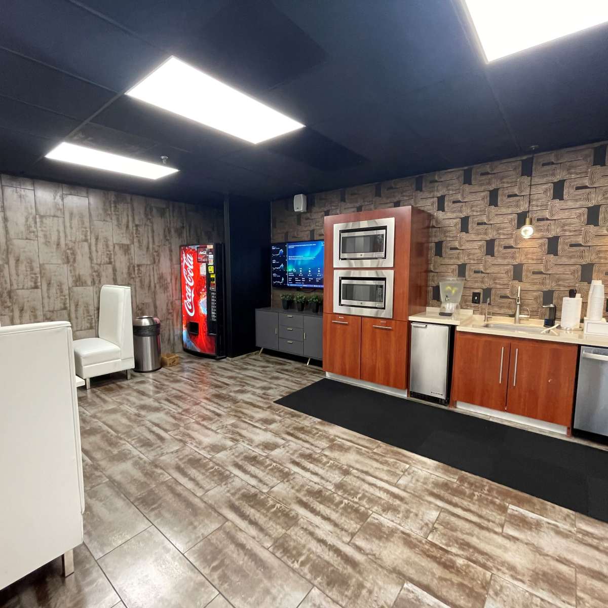 A modern kitchen space with stainless steel appliances, a vending machine, a television on the wall, and seating in a corner.