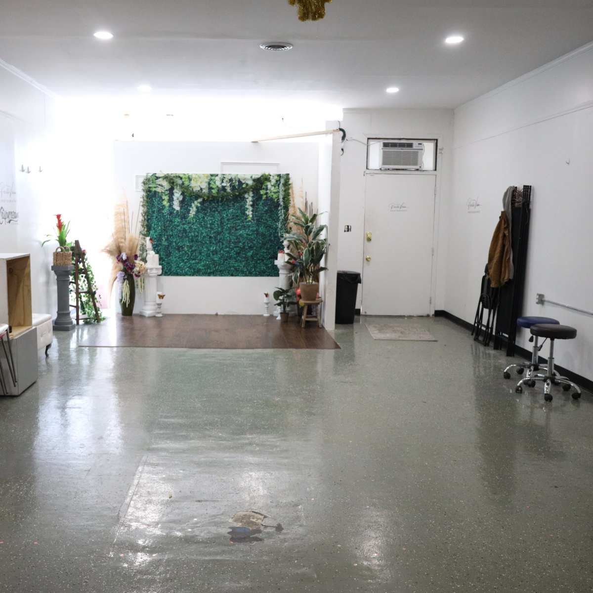 An empty room features a decorated wall of greenery, a small table, and several plants arranged around a polished floor.