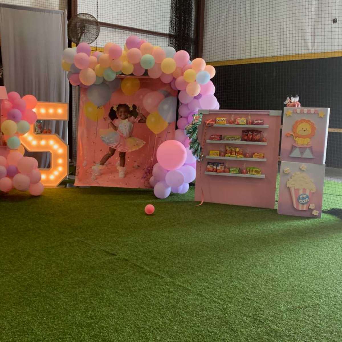 A decorated indoor party area featuring a balloon arch, a photo backdrop, a candy cart, and a shelf with snacks on a faux grass floor.