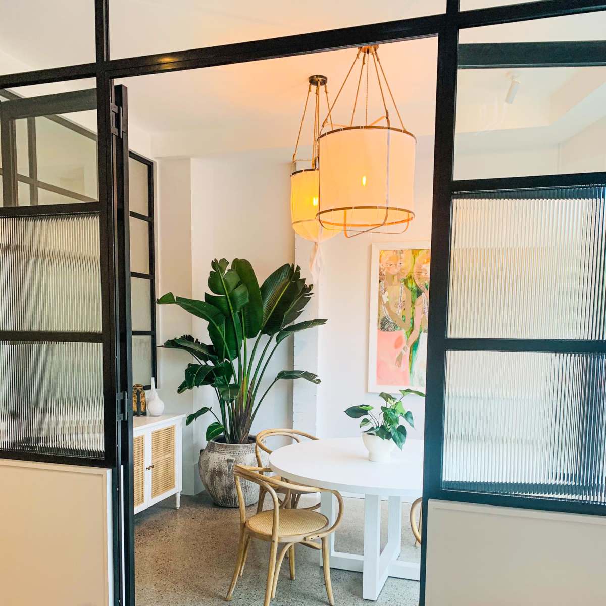 A bright, modern dining area features a round table surrounded by chairs, with large plants and decorative art displayed on the walls.