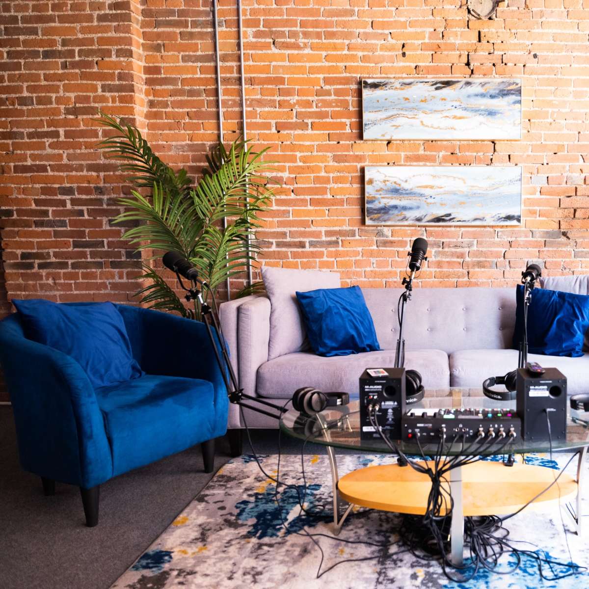 The scene shows a modern podcasting setup with a gray couch and blue chairs, featuring microphones and headphones on a glass table in front of a brick wall.
