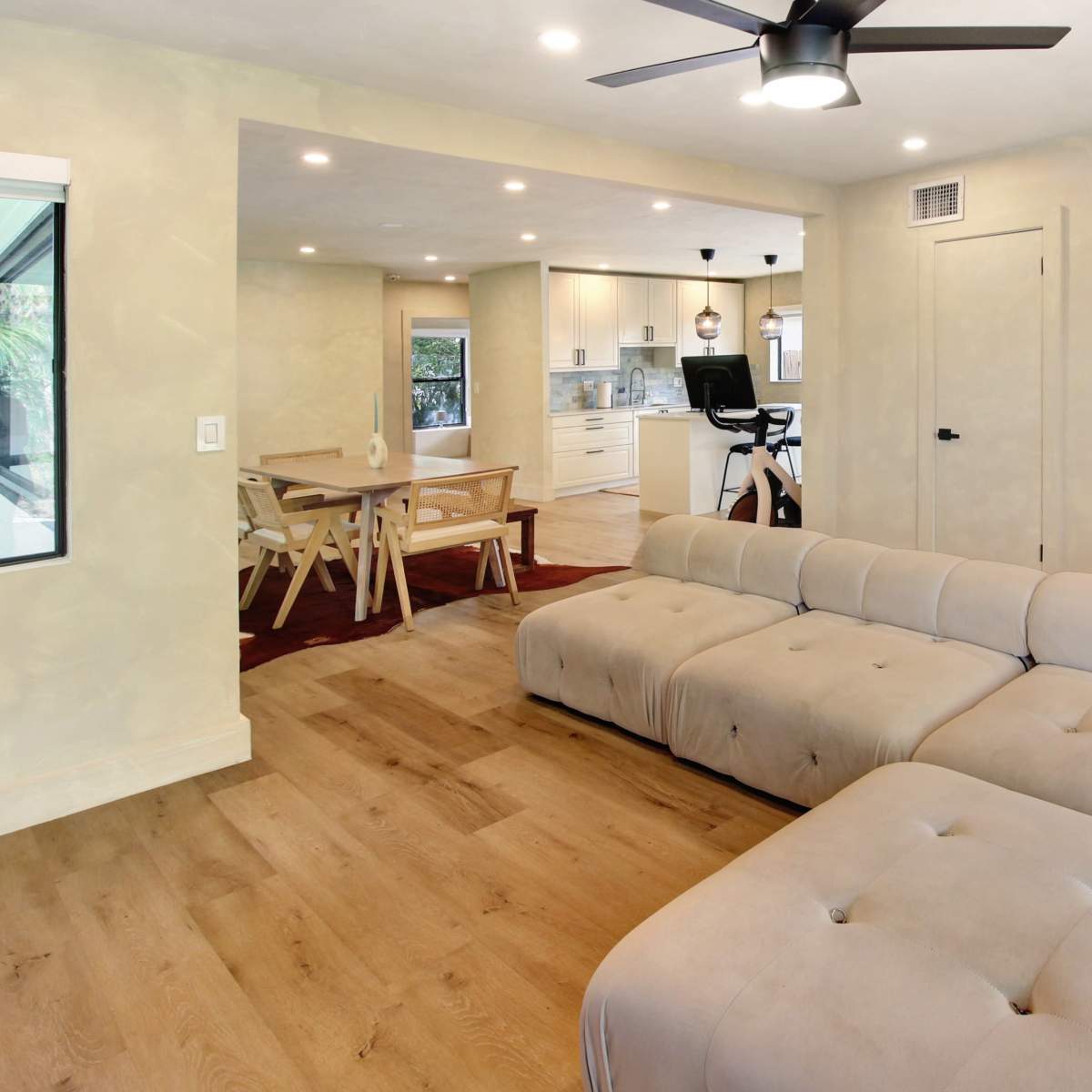 A modern living room featuring a large beige sectional sofa, a wooden dining table, and an open kitchen area with bright lighting.