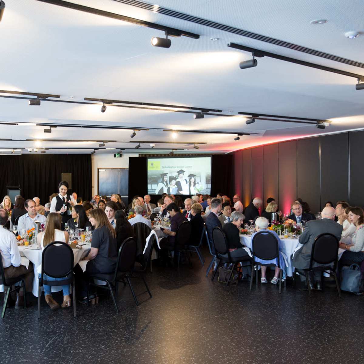 A large group of people is seated around multiple tables in a spacious, well-lit event hall, enjoying a meal together.