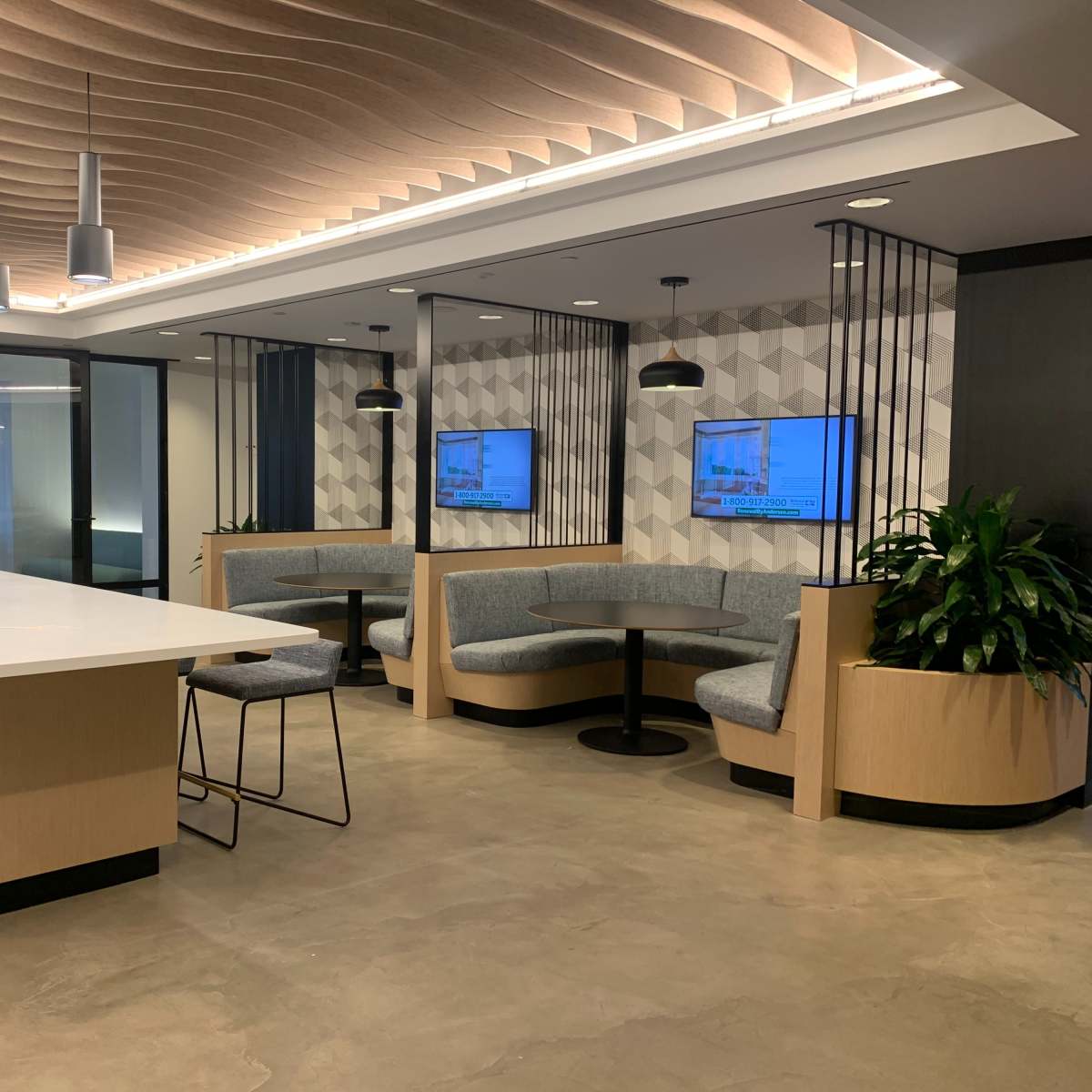 A modern office reception area with a central island, sectional seating, and two wall-mounted screens.