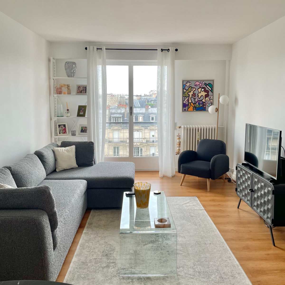 A modern living room features a gray sectional sofa, a glass coffee table, and a black armchair, with large windows providing a view of city rooftops.