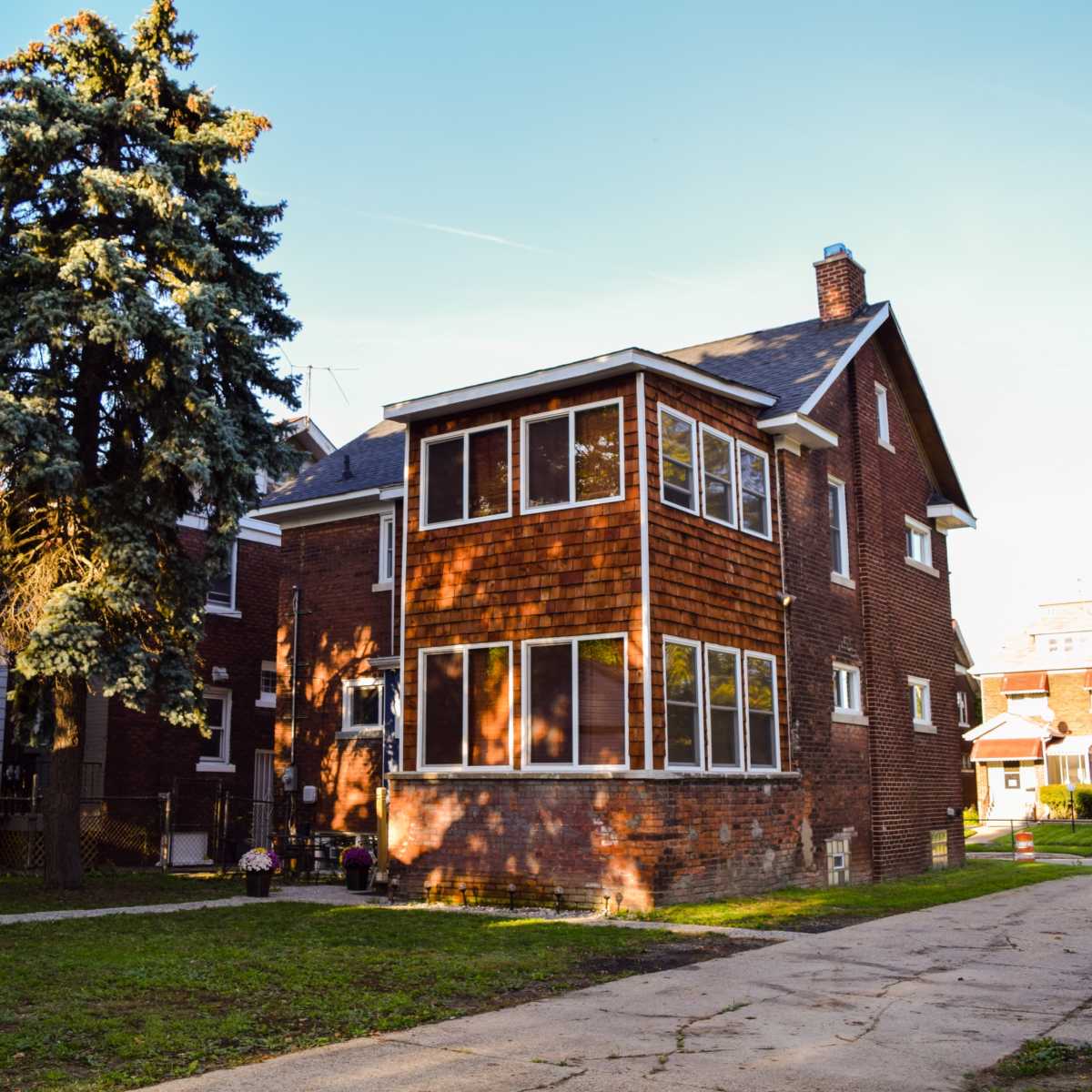 A multi-story brick house with large windows and a tree in the front yard, set on a residential street.