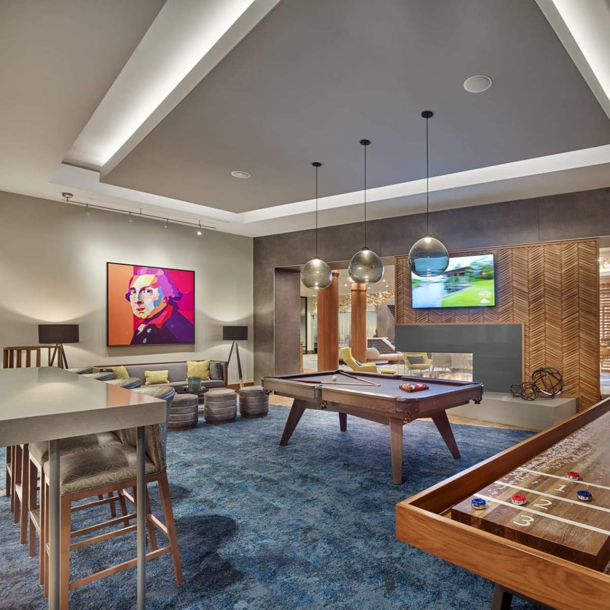 A modern game room with a pool table, shuffleboard, and an illuminated ceiling, featuring a colorful wall art piece and a television in the background.