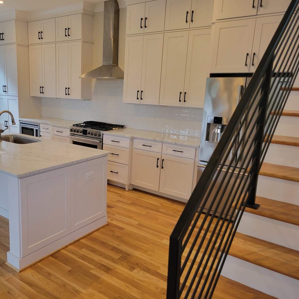 A modern kitchen with white cabinetry, a large island, stainless steel appliances, and a wooden staircase in the background.