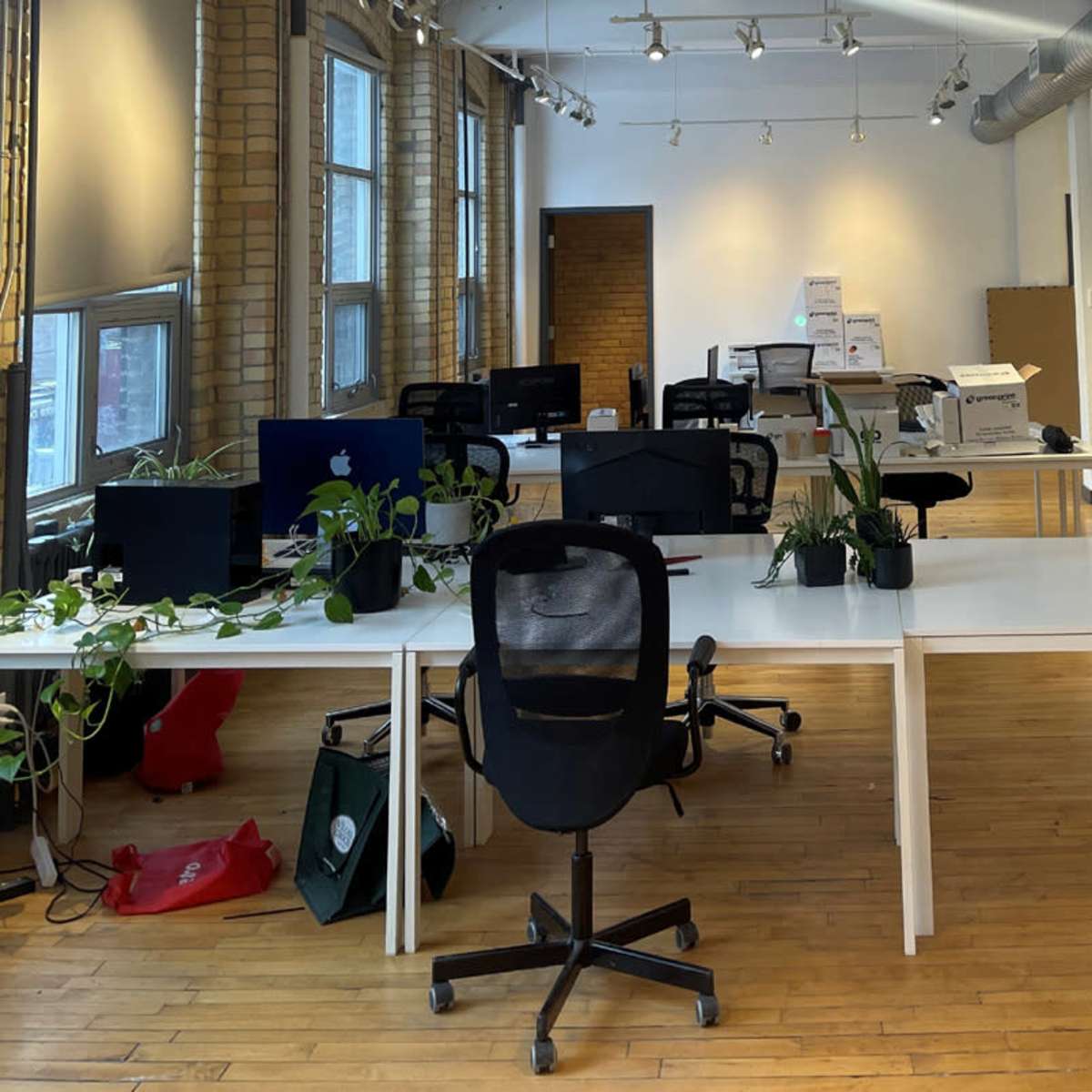 An office space with several white desks, rolling chairs, and minimal decorations, illuminated by overhead lighting.