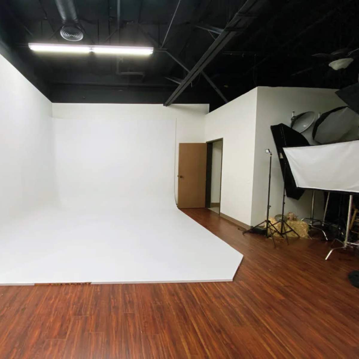 A spacious photography studio featuring a seamless white backdrop, wooden flooring, and several lighting setups on one side.
