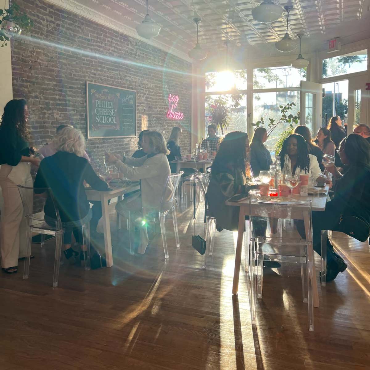 The scene shows a lively restaurant with patrons seated at tables, enjoying drinks and conversation, as sunlight streams through large windows.
