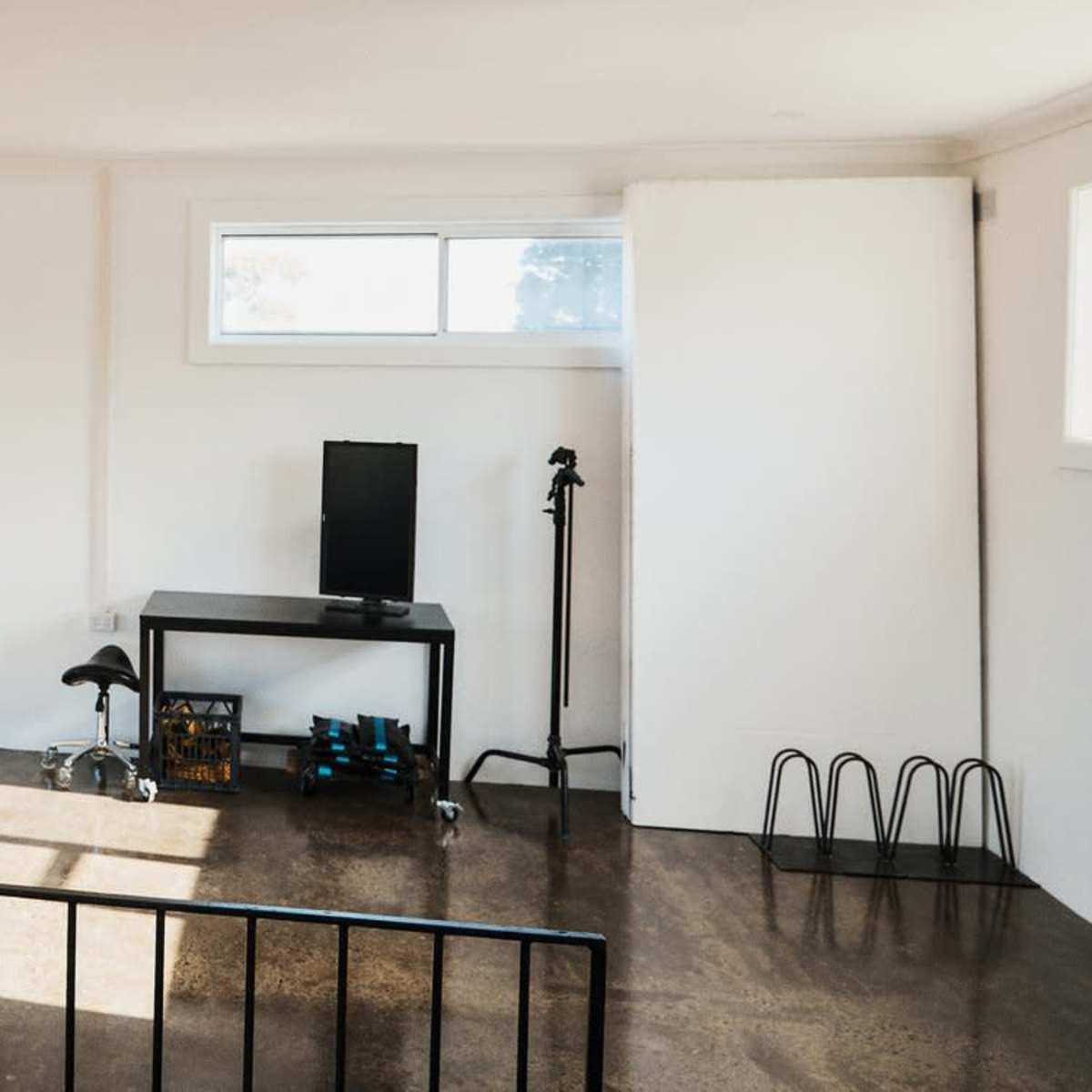 A minimalistic room with two windows, a black table with a monitor, a chair, and a rack for weights against a white wall.