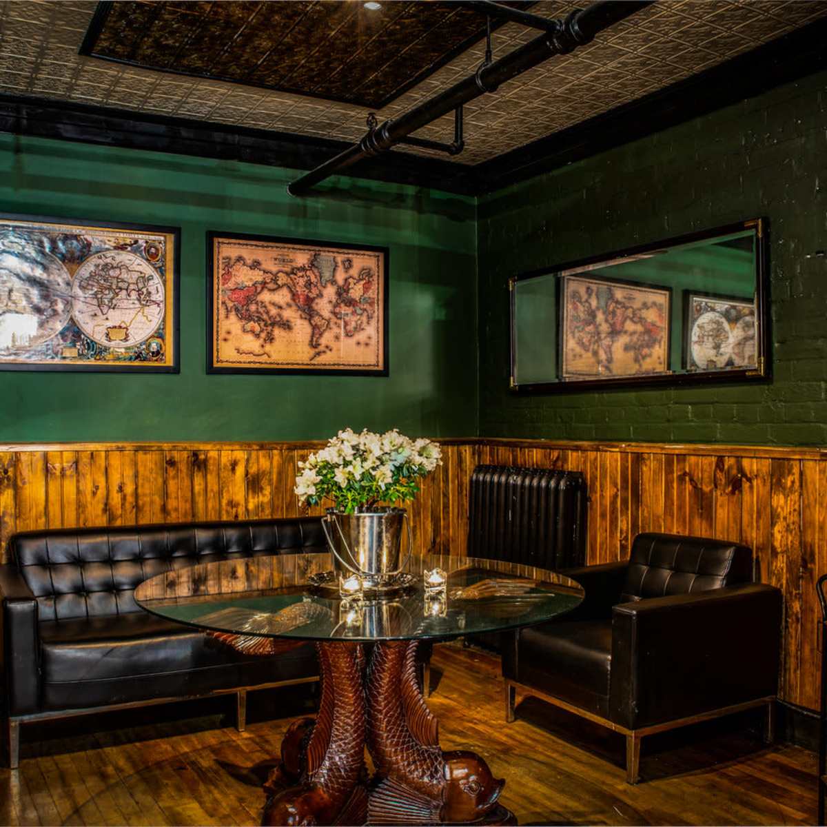 A cozy lounge area with dark wood paneling, a glass table, and framed maps on the green walls.