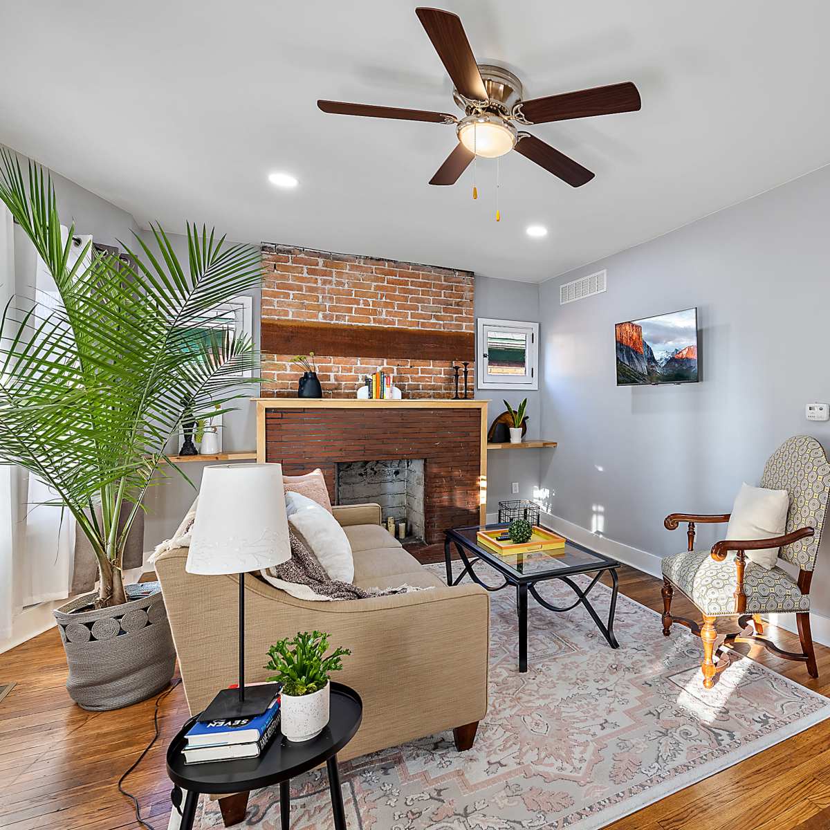 A cozy living room with a ceiling fan, a brick fireplace, and a mix of modern and traditional furnishings.