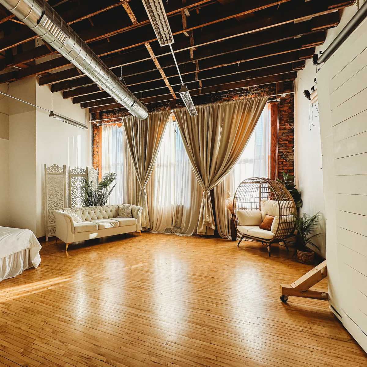 A spacious room with wooden floors, large windows covered by drapes, a bed, a sofa, and a wicker chair.