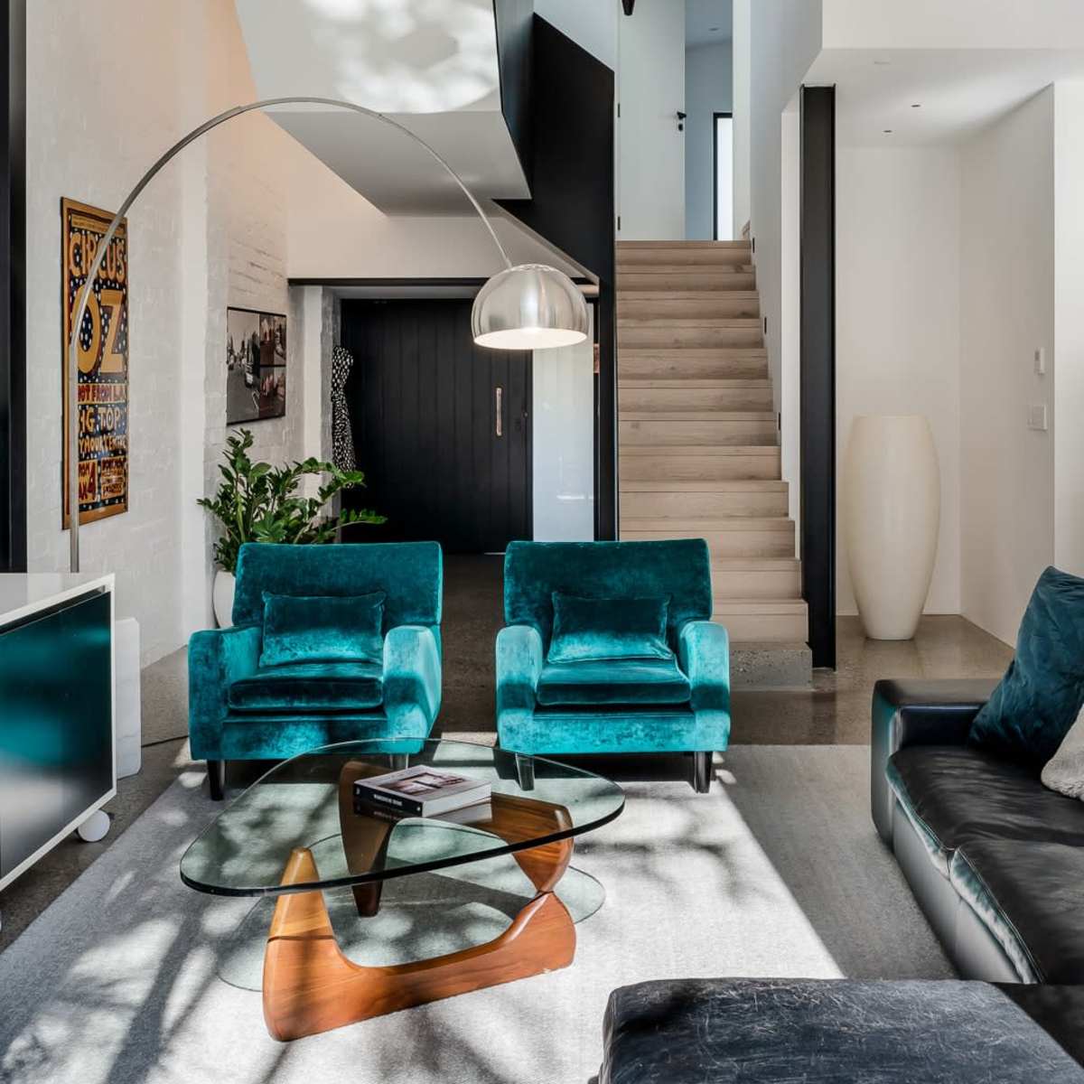 A modern living room featuring two teal armchairs, a glass coffee table, and a staircase leading to an upper level.