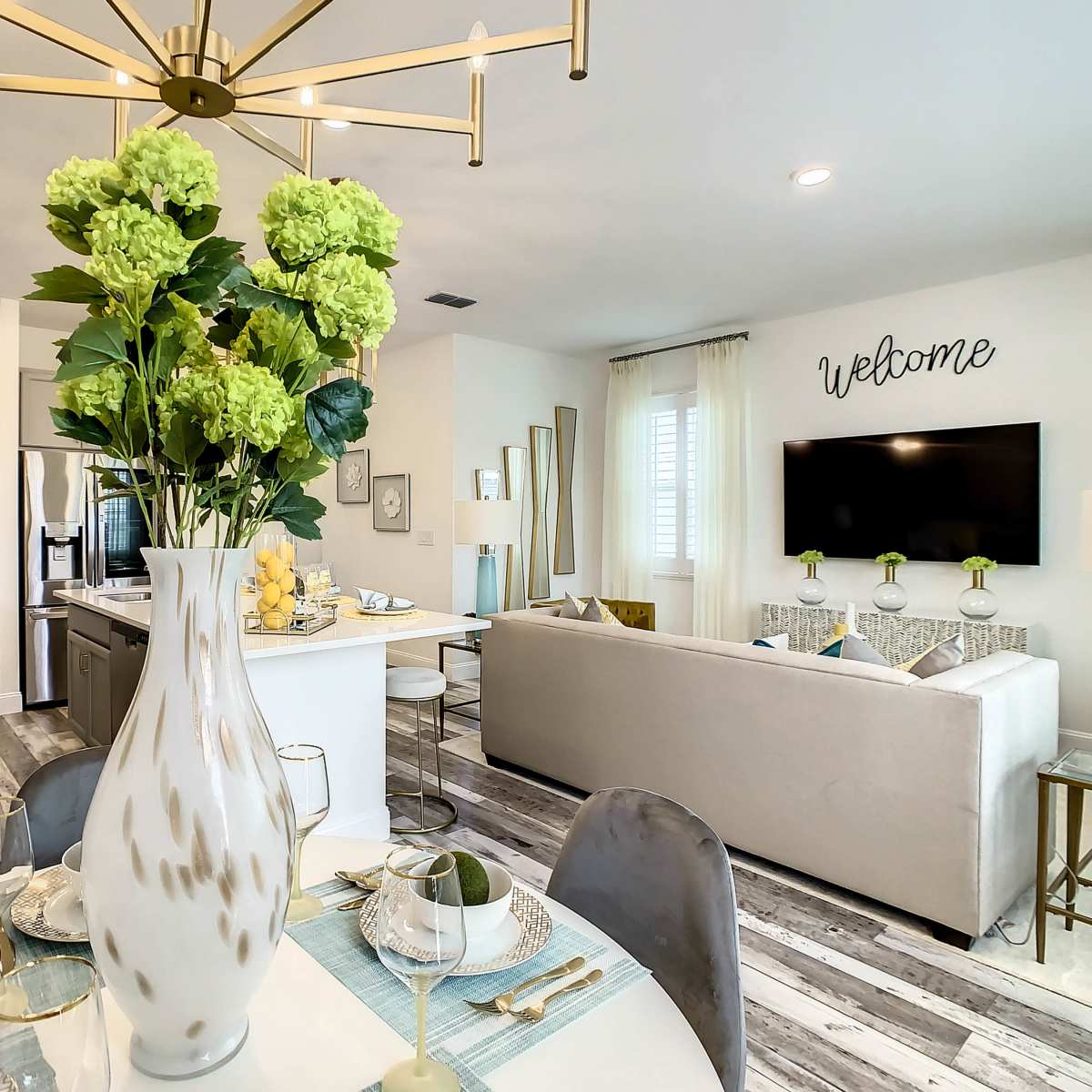A modern living space features a dining table with a large vase of green flowers, an adjacent kitchen area, and a cozy sitting area with a television.