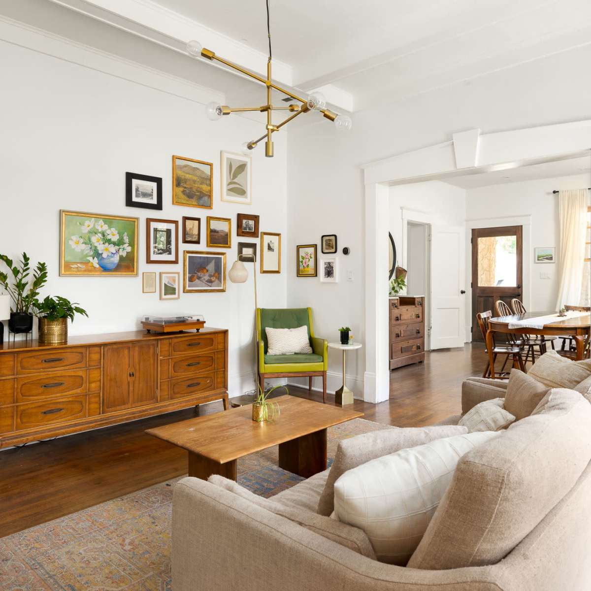 A cozy living room features a beige sofa, a wooden coffee table, and a sideboard adorned with framed artwork and plants, with an adjacent dining area visible in the background.