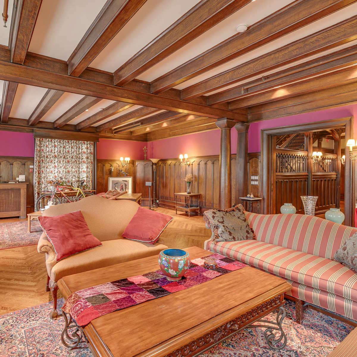The room features wooden beams on the ceiling, a mix of furniture including a striped sofa and a coffee table, and decor elements such as plants and a patterned area rug.