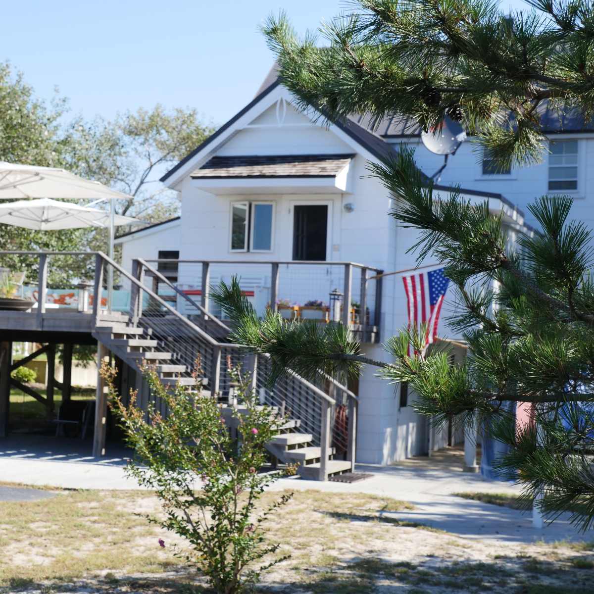 A two-story house with a deck and stairs leads to an outdoor seating area, featuring umbrellas and an American flag, surrounded by greenery.