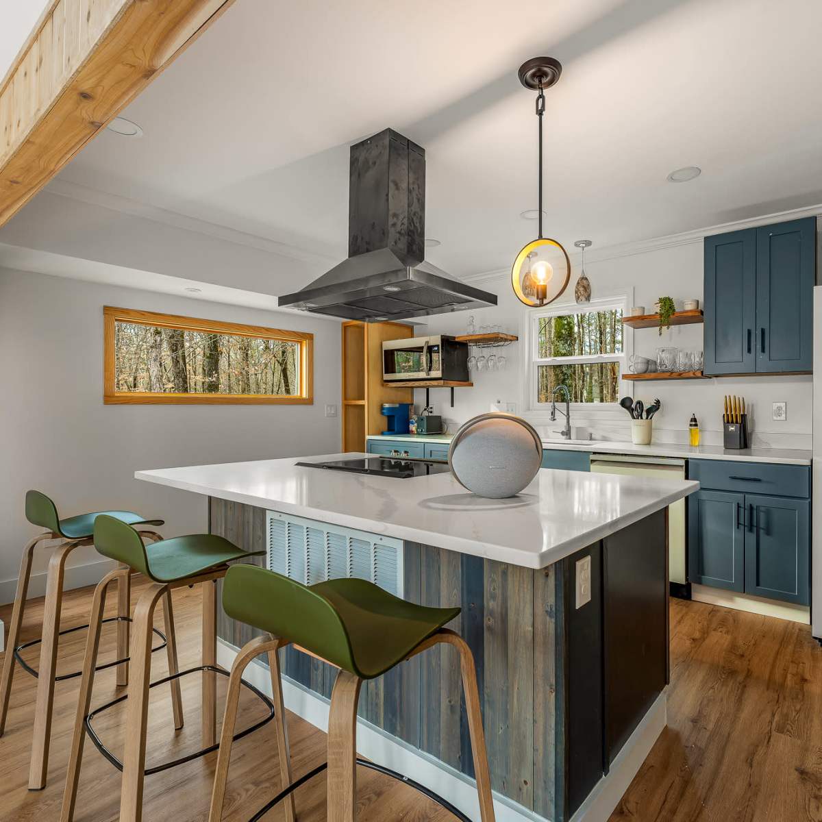 The kitchen features a central island with bar seating, a metal range hood, and blue cabinets against a backdrop of large windows.