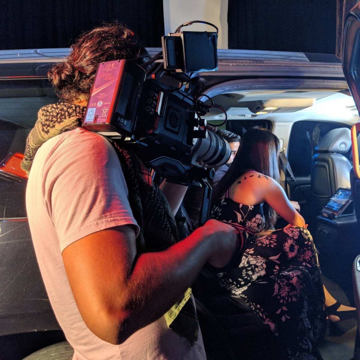A camera operator films a woman sitting inside a vehicle, reviewing content on a device.