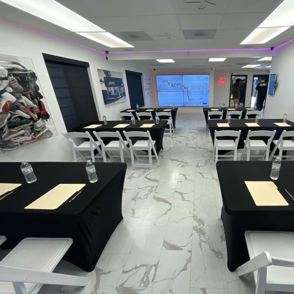 A conference room is set up with black tables and white chairs, featuring a large screen displaying data charts and graphs on one wall.