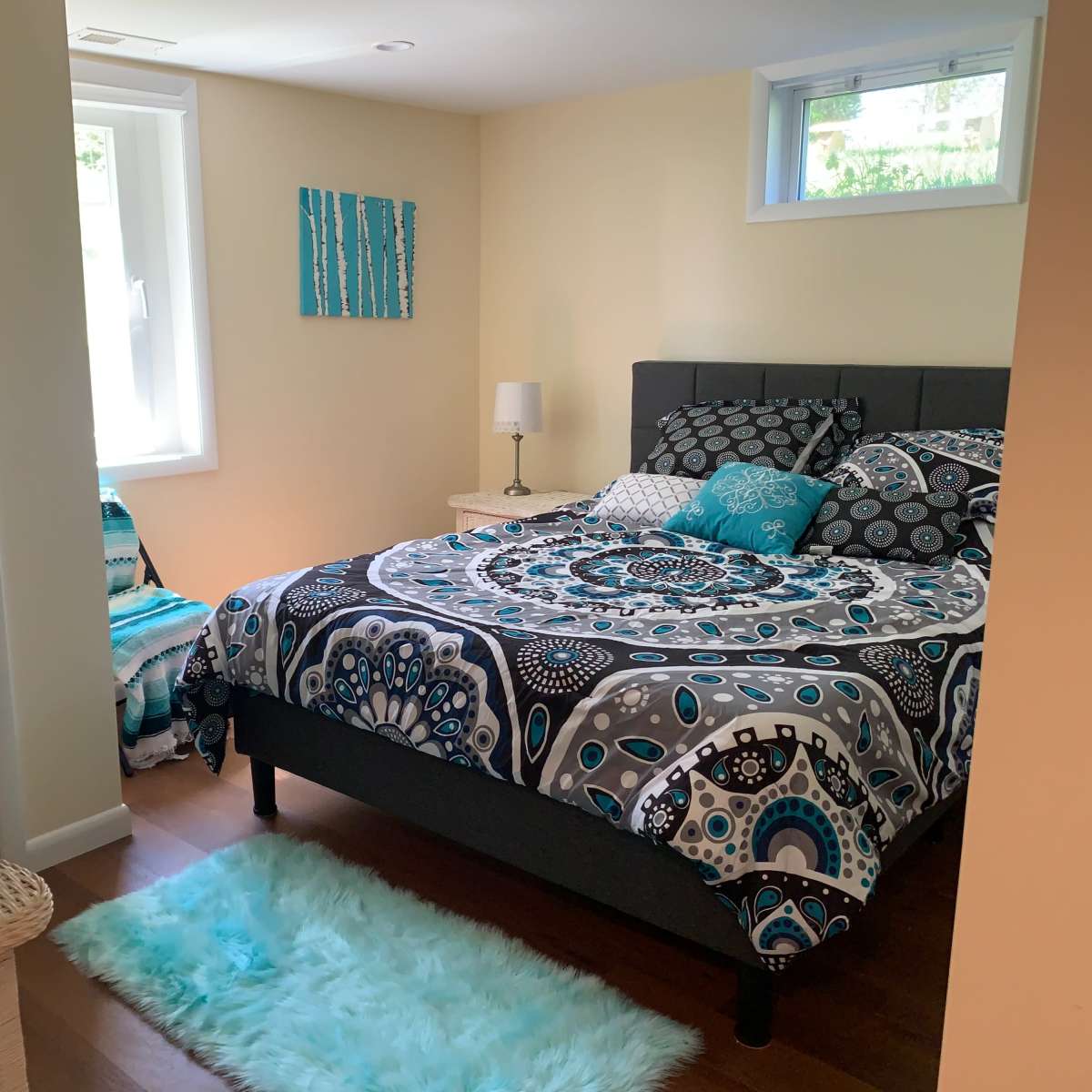 A small bedroom features a bed with a patterned blue and gray comforter, a nightstand with a lamp, and a chair, while towels are neatly stacked on a wicker basket.