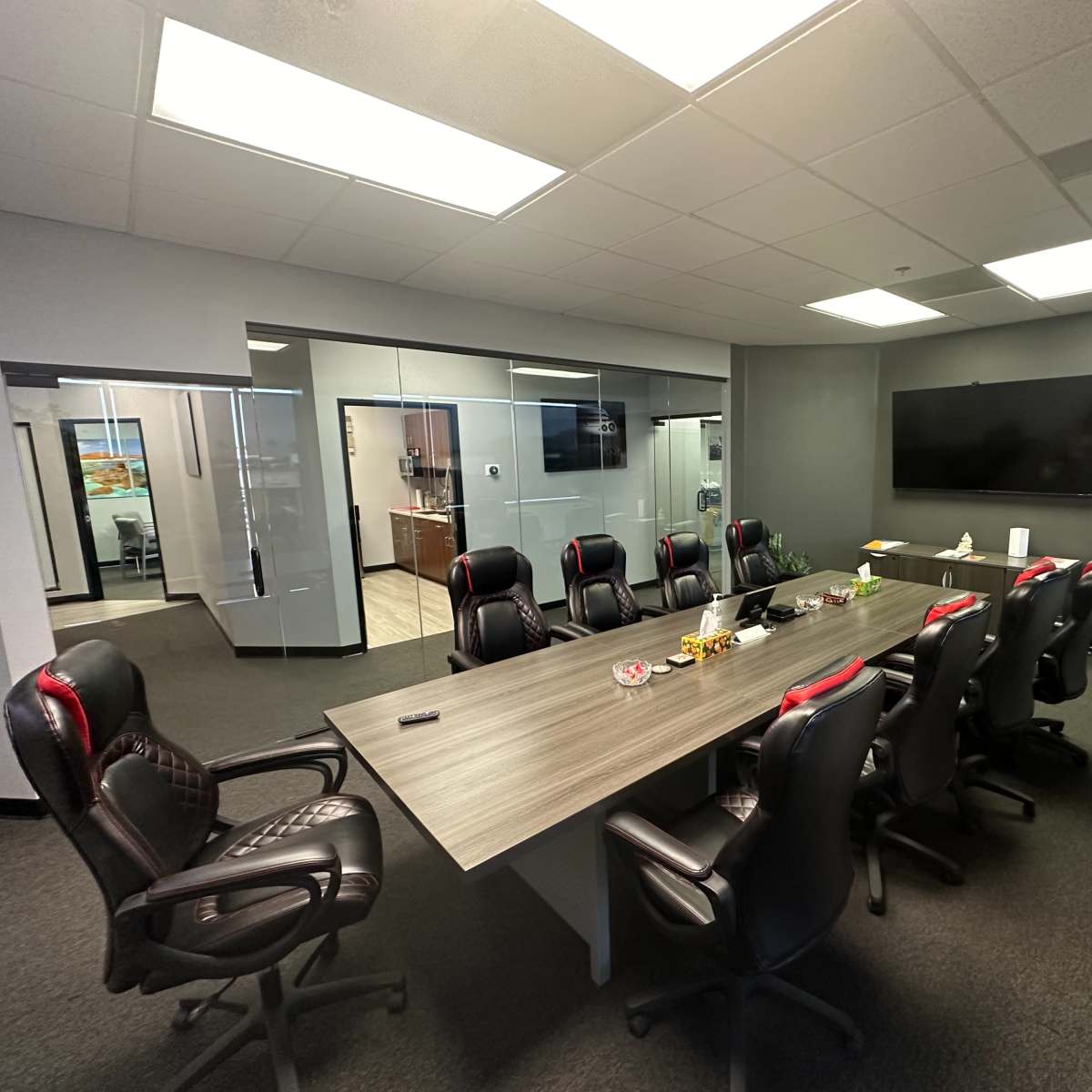 A modern conference room with a long table and black office chairs arranged around it, adjacent to glass-walled offices.