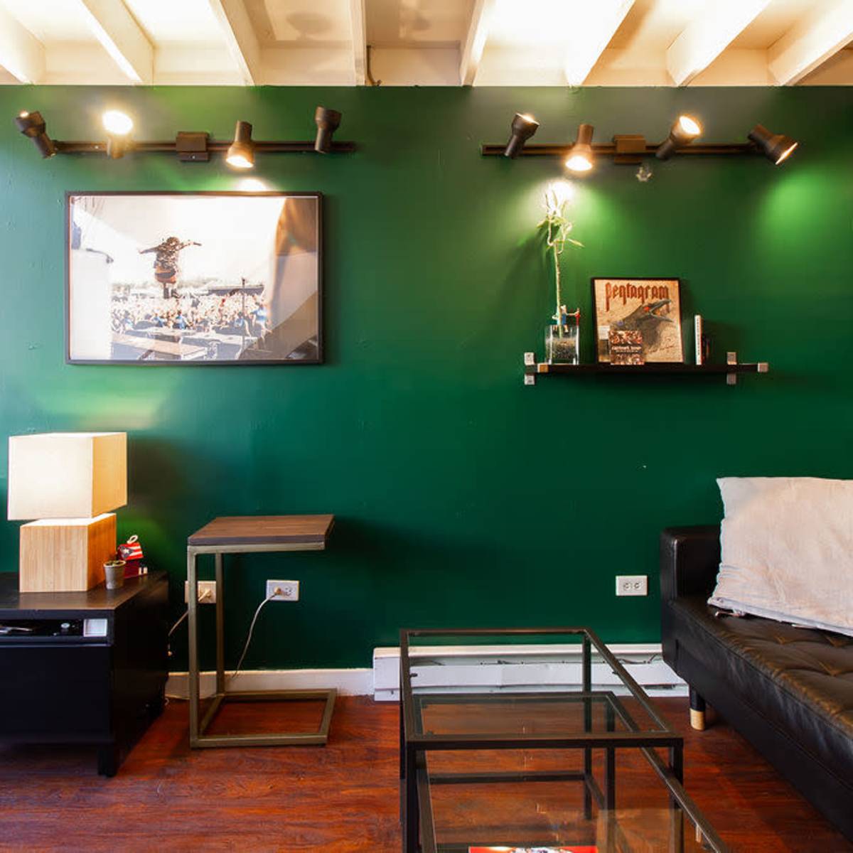 A modern living room with a green accent wall, featuring a black leather sofa, a glass coffee table, and a wall-mounted shelf with decorative items.