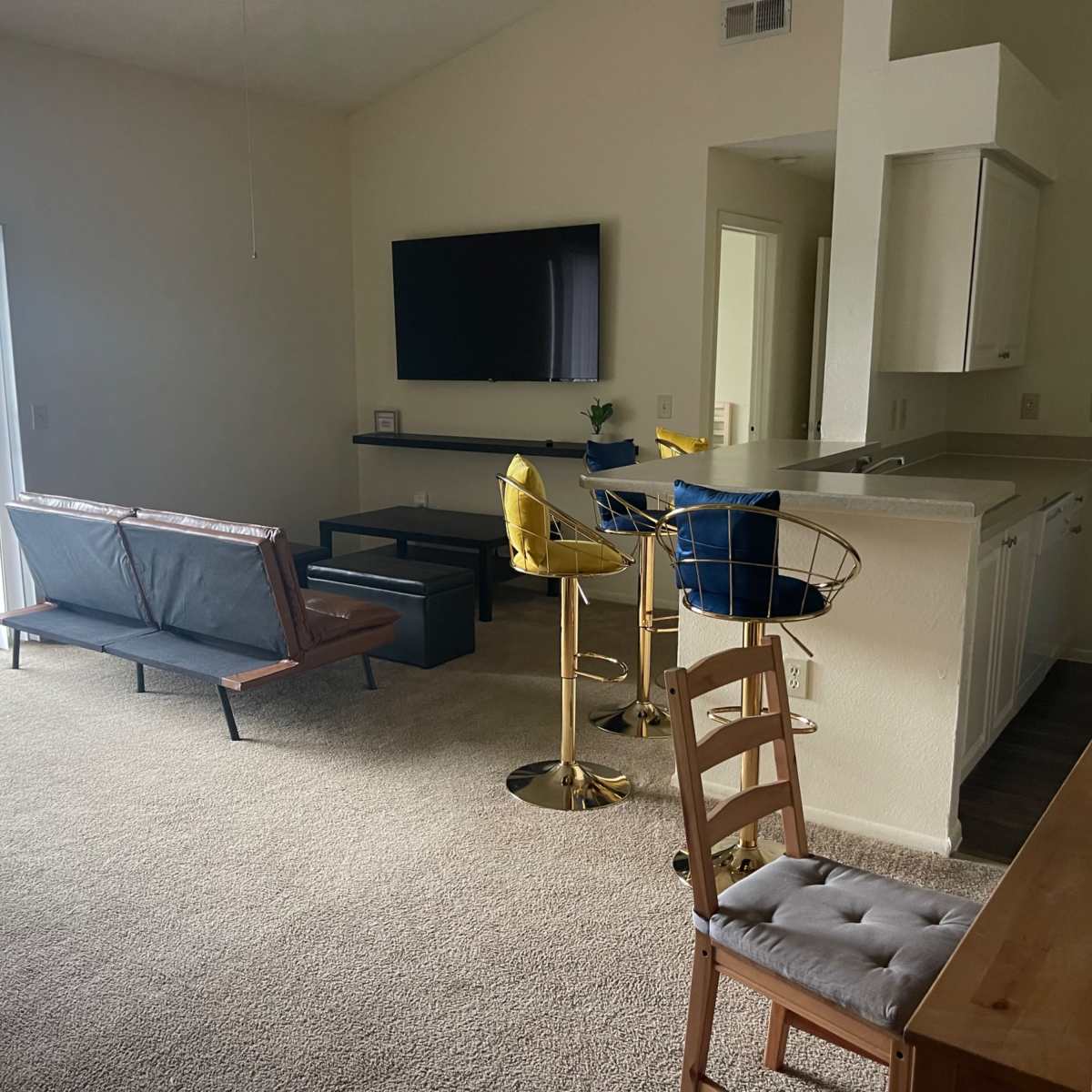 The living space features a blend of seating options, including a sleek couch, bar stools, and a dining chair, all arranged in a sunlit room with a television and a sliding door leading to a balcony.