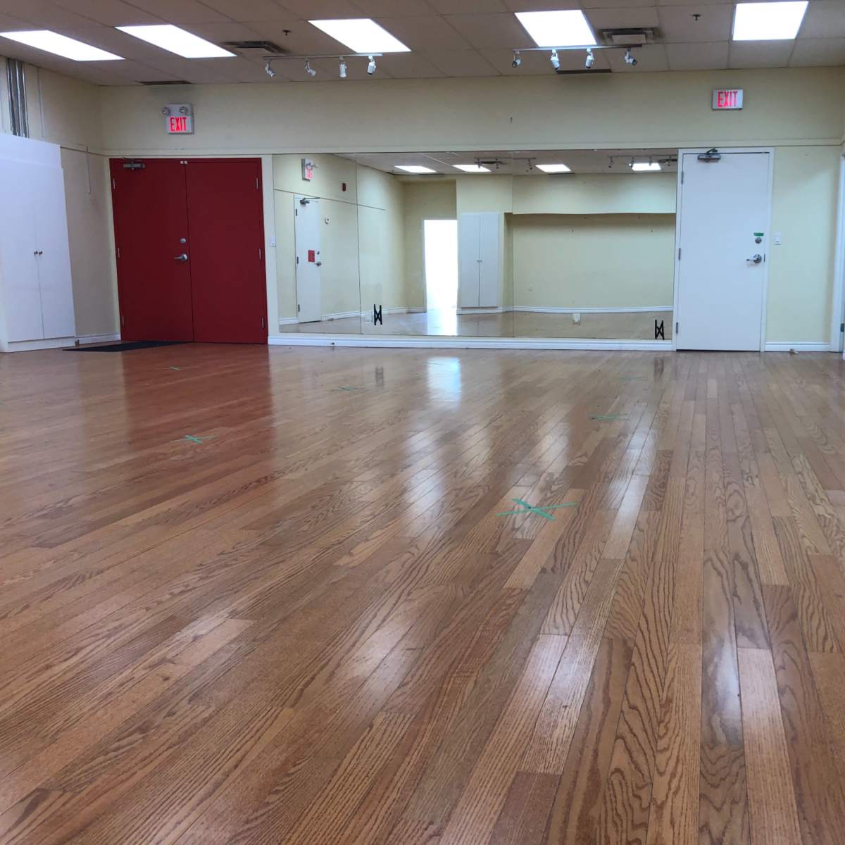 A brightly lit dance studio with polished wooden floors, two red doors, and large mirrors on one wall.