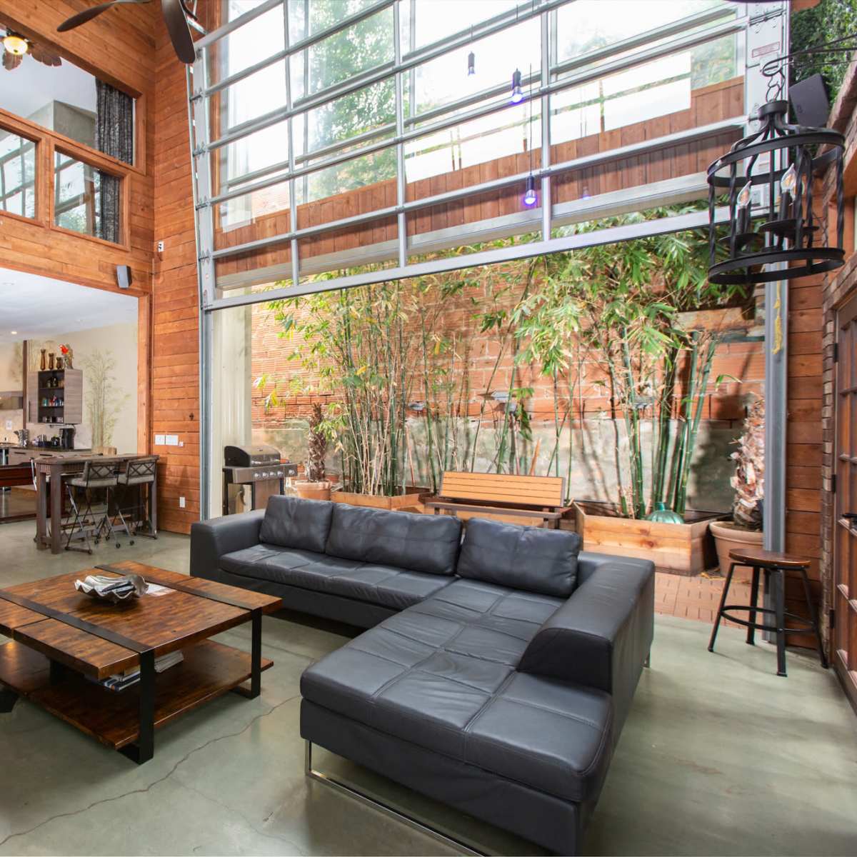 A modern living room with wooden walls, a high ceiling, and large windows overlooking a bamboo garden, featuring leather sofas and a wooden coffee table in the center.